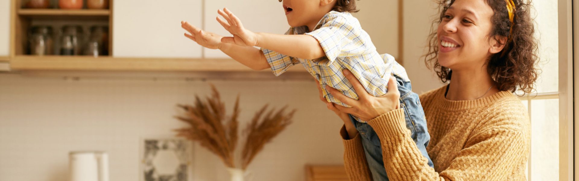 Portrait of cute young Latin woman in sweater sitting on wndowsill holding her two year old son who is reaching out hands as if flying. Happy mom and child playing in cozy kitchen interior